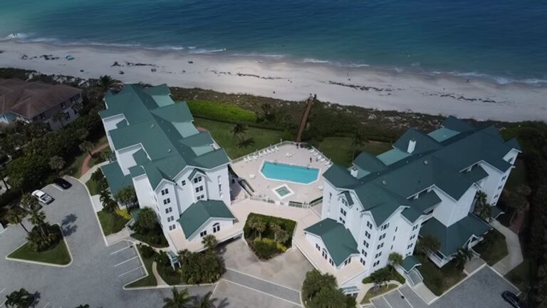 Aerial view of a coastal resort with a pool, next to a sandy beach with waves.