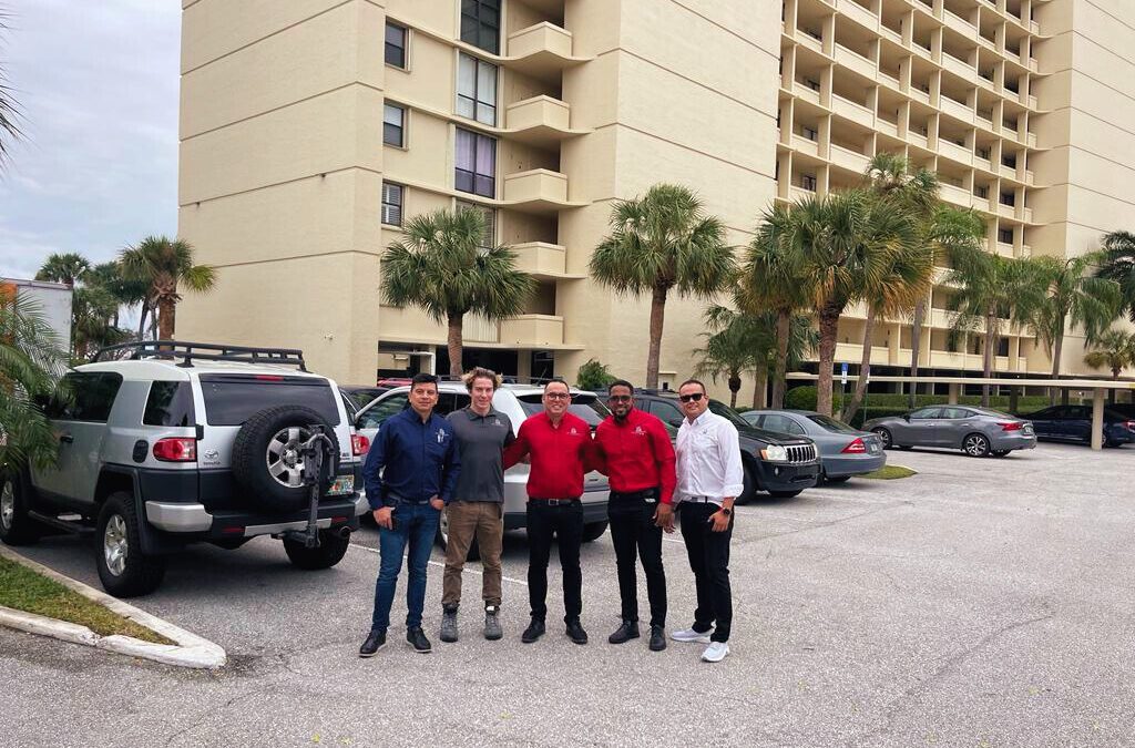 Group of people standing in a parking lot with cars and palm trees nearby.