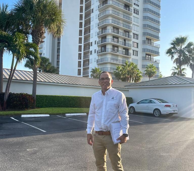 Man in business attire holding paperwork, standing in a parking lot with a high-rise building behind.